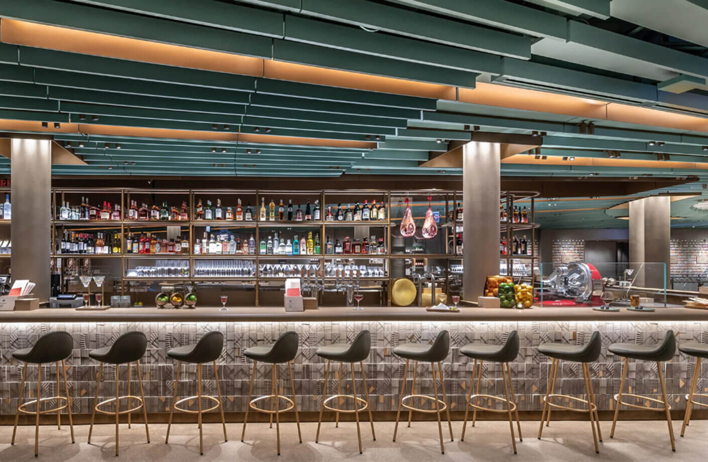 Stylish bar featuring geometric ceiling details, a sleek counter, and a variety of illuminated shelves showcasing an extensive drink selection.