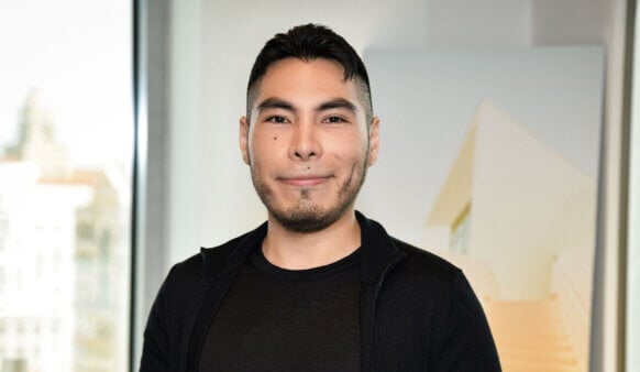 A man with short dark hair and a trimmed beard smiles, wearing a black shirt and sweater, standing in an office with a soft background.