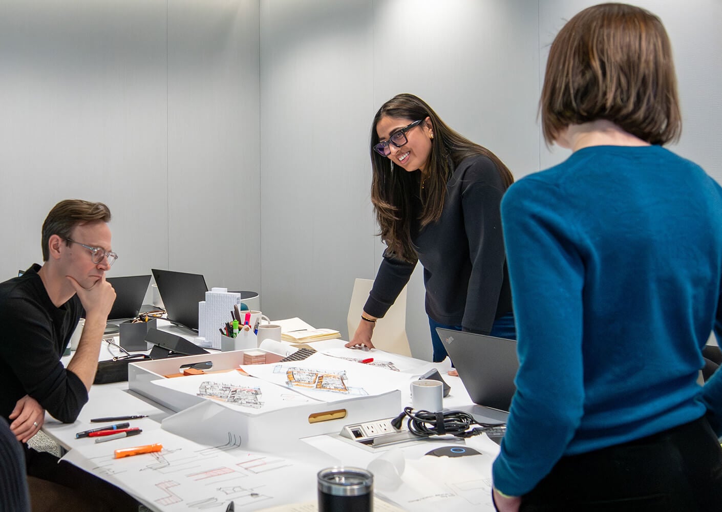 Ananta Sodhi discussing with teammates at a table covered with drawings and notes. Office setting with modern design elements.