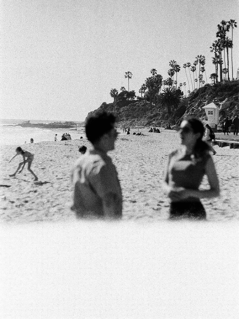 Black and white film photo of people talking on a beach, with palm trees and a rocky coast in the background. Others are enjoying the sandy shoreline.