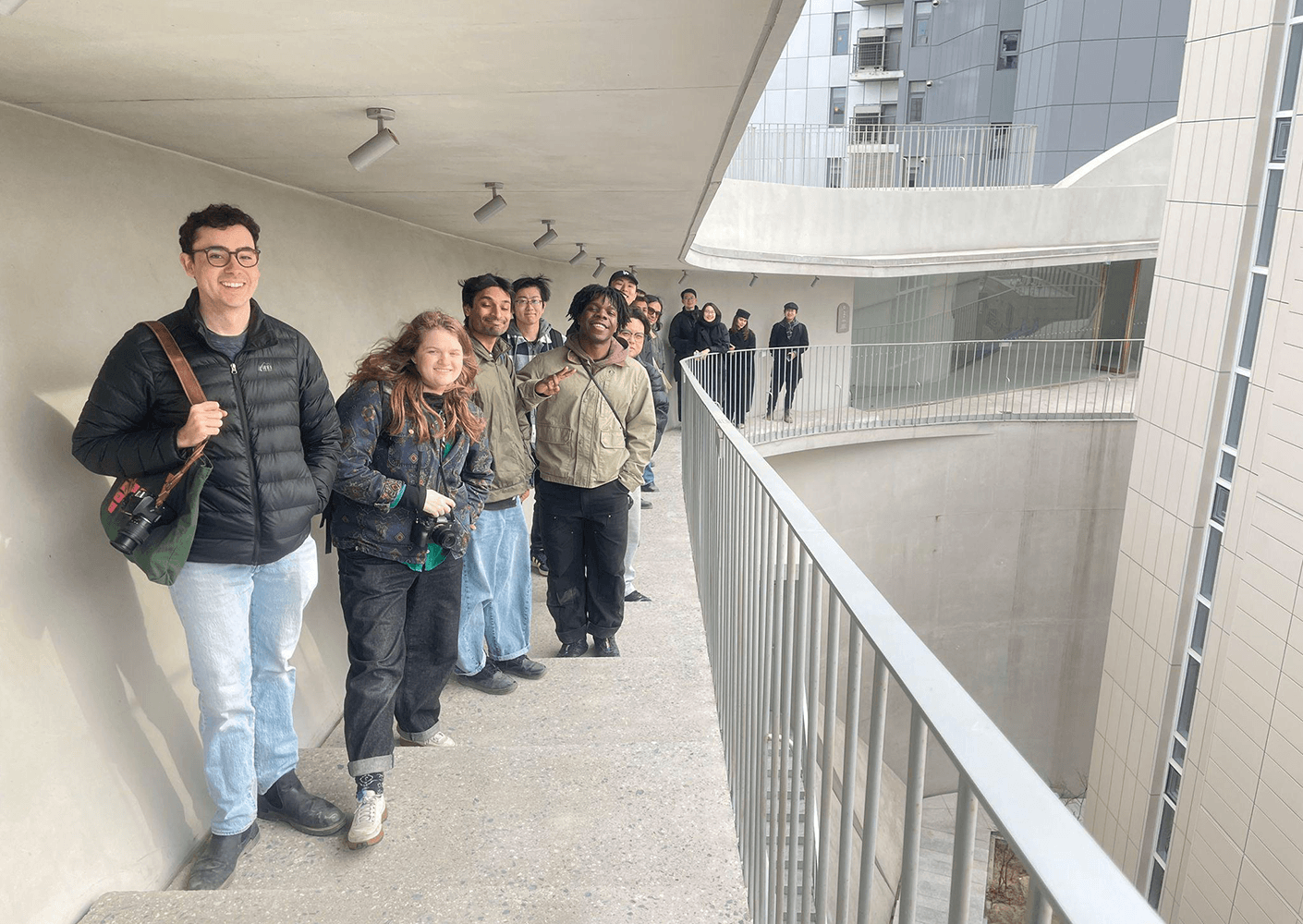 A group of students stands on a curved balcony, smiling and chatting, with a modern architectural structure in the background.