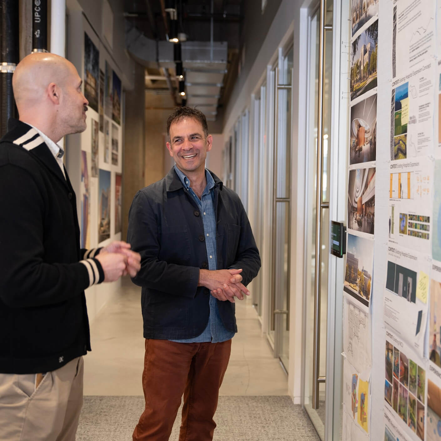 Gilad and a team member converse in a hallway, examining architectural boards on the wall. The space features large windows and a clean design.