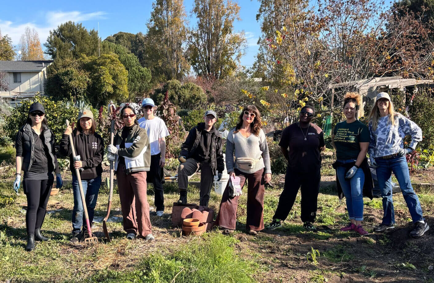 Group of people in a community garden, holding gardening tools and standing among plants, with trees and a house in the background.