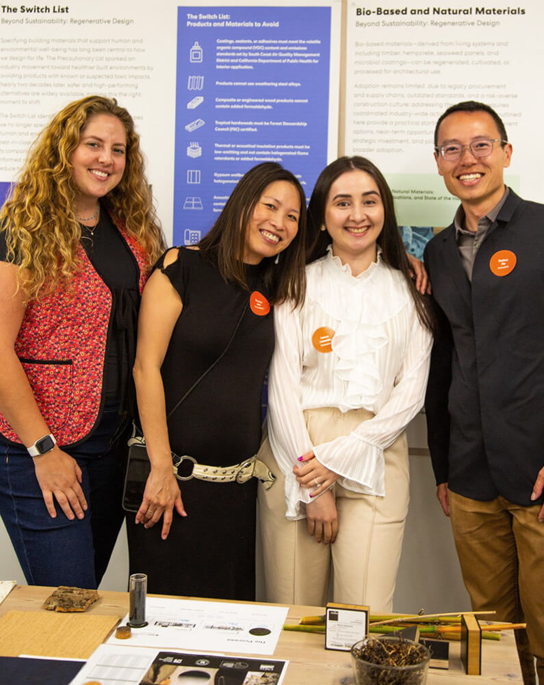 Four individuals pose together in a workspace, showcasing design materials and sustainability projects, with visual information displayed in the background.