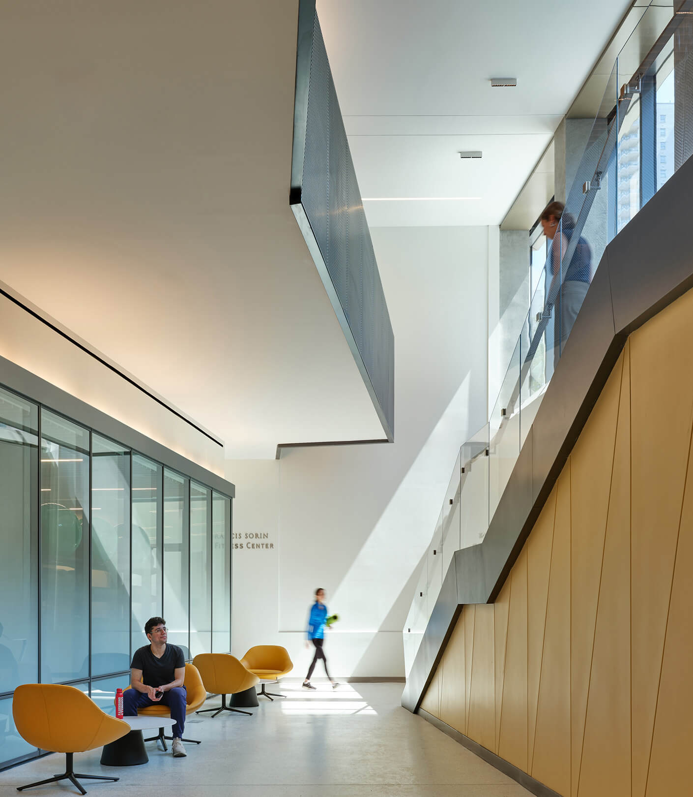 Interior view of a fitness center with seating, a staircase, and large windows allowing natural light. People are interacting in the space.