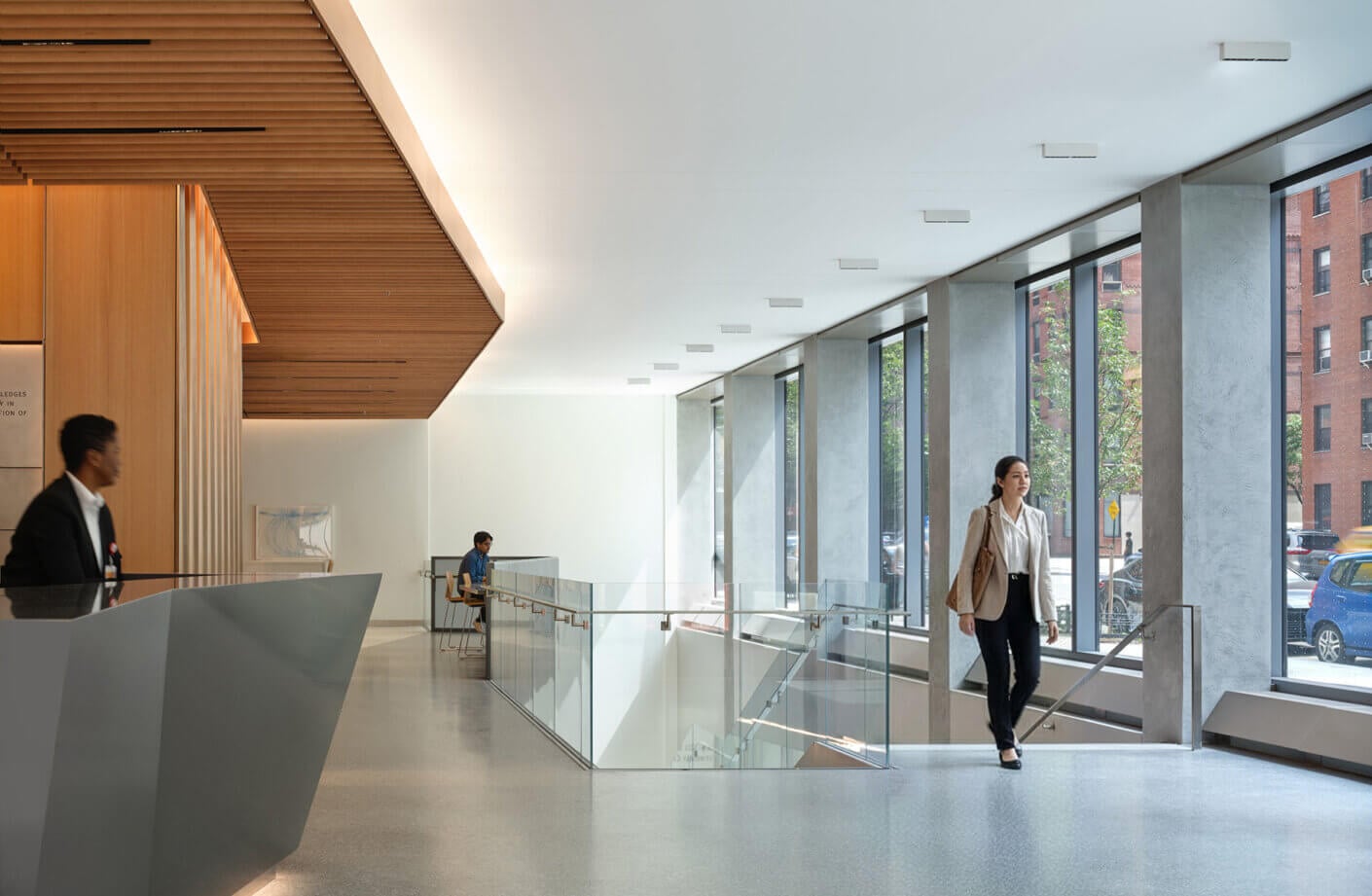 Interior view of a spacious lobby with large windows, glass railings, a concierge, and individuals engaging inside the space.