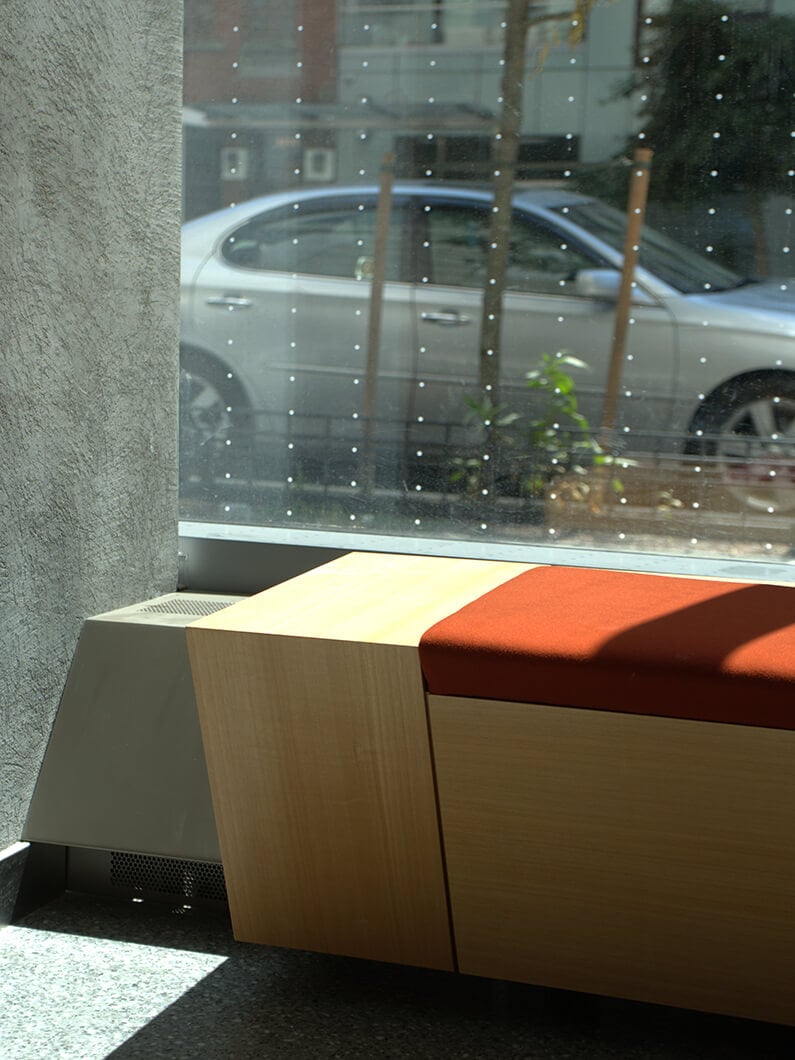 Interior seating area featuring a simple wooden bench with an orange cushion by a large window, overlooking a street with a parked car.
