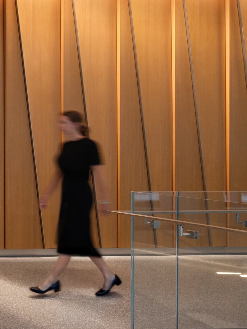 A person walks past a wooden wall with vertical lines, alongside a glass railing in a well-lit interior space.