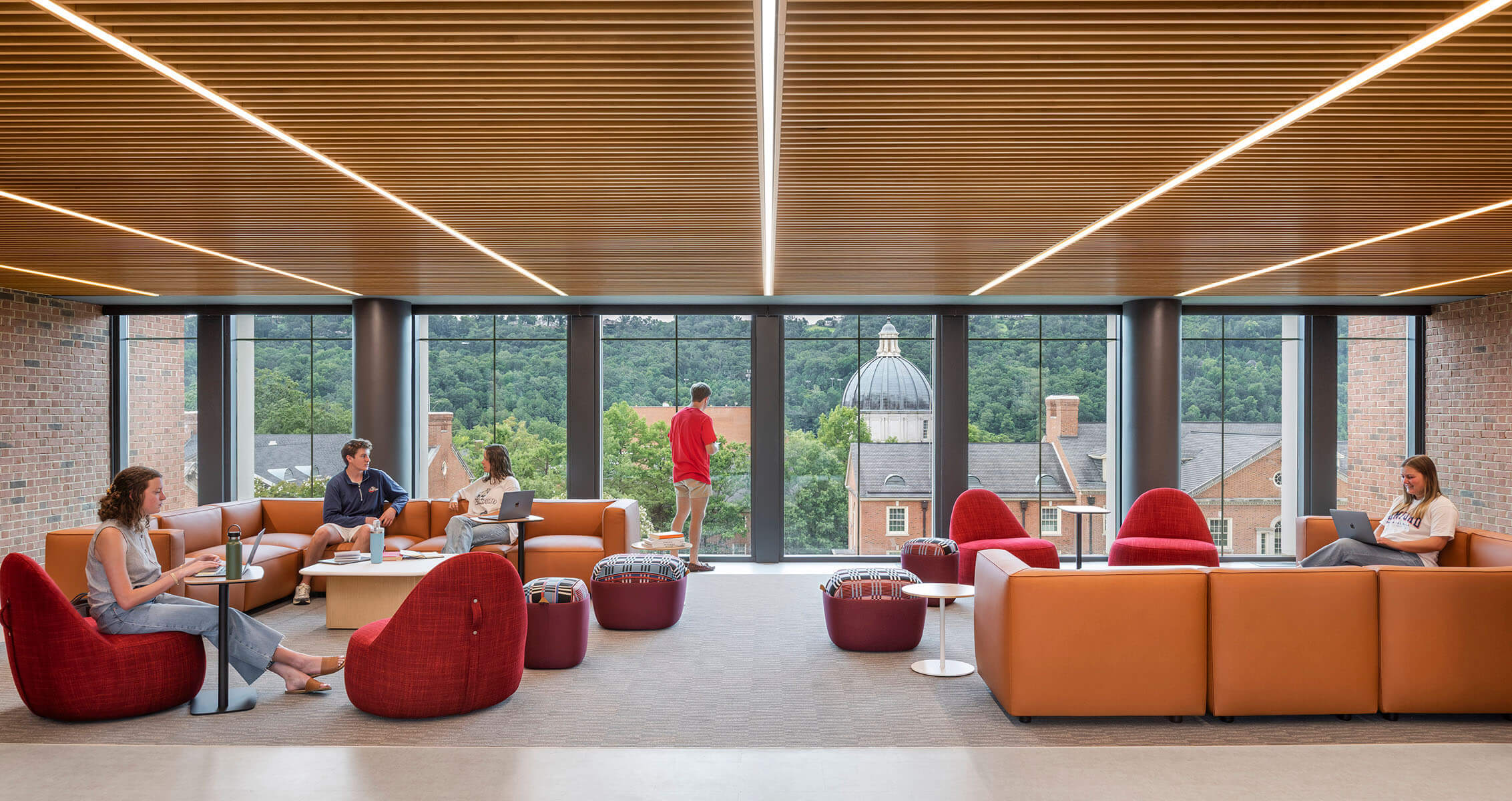 Students working and relaxing in a spacious lounge with large windows and comfortable seating, overlooking a green outdoor view.