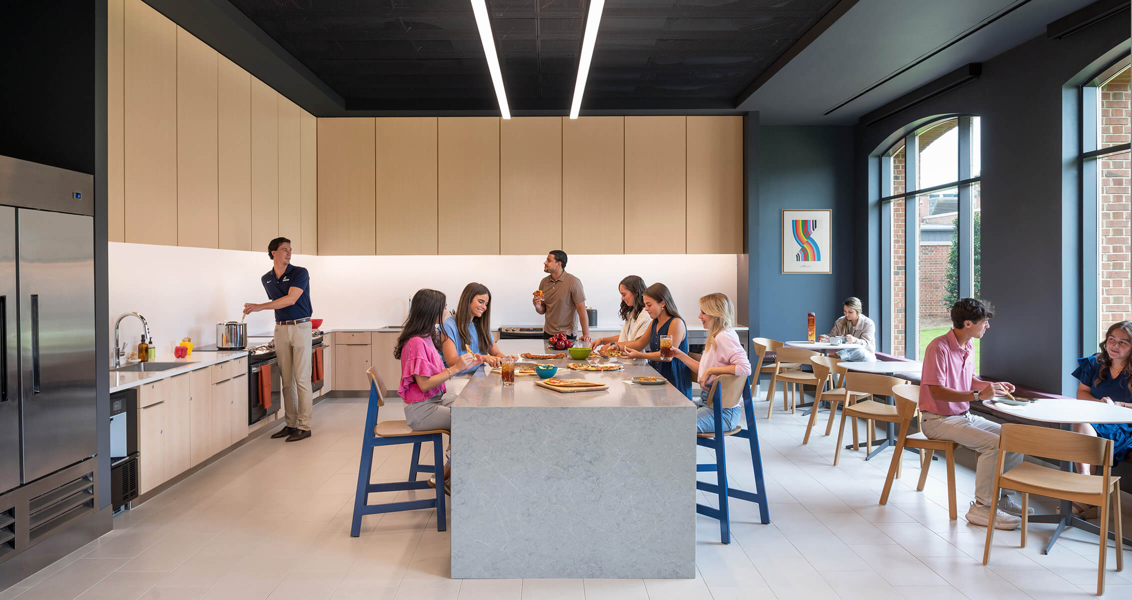Group of people socializing in a kitchen and dining area, featuring a large island, modern appliances, and ample natural light.