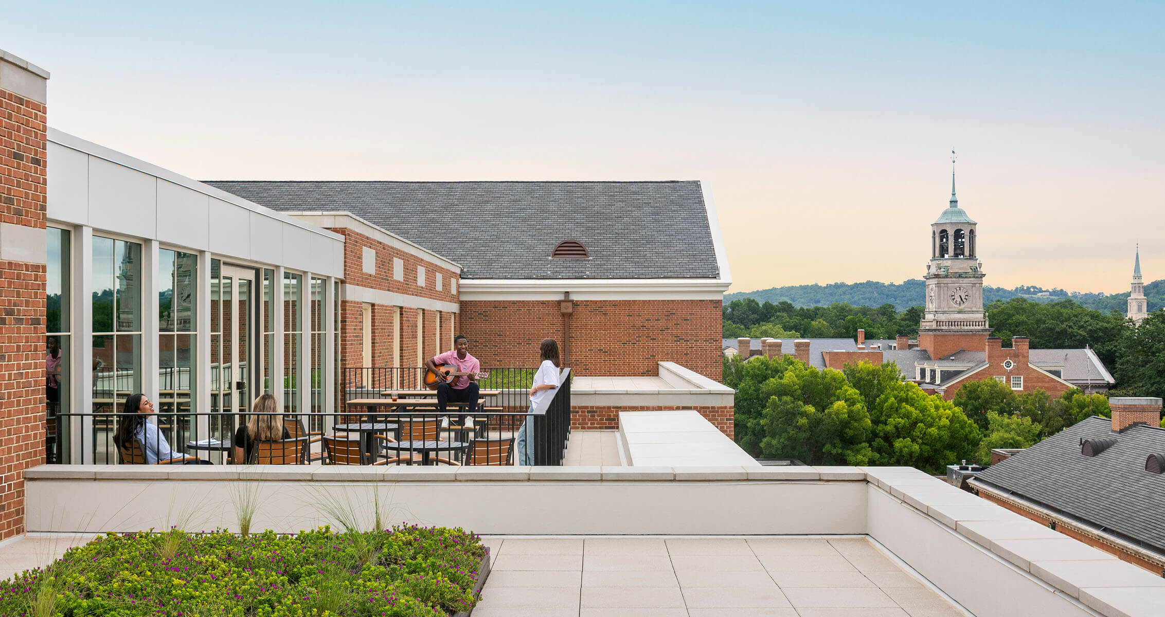 People relaxing on a spacious rooftop terrace with seating and green landscaping, overlooking a skyline with church steeples and trees.