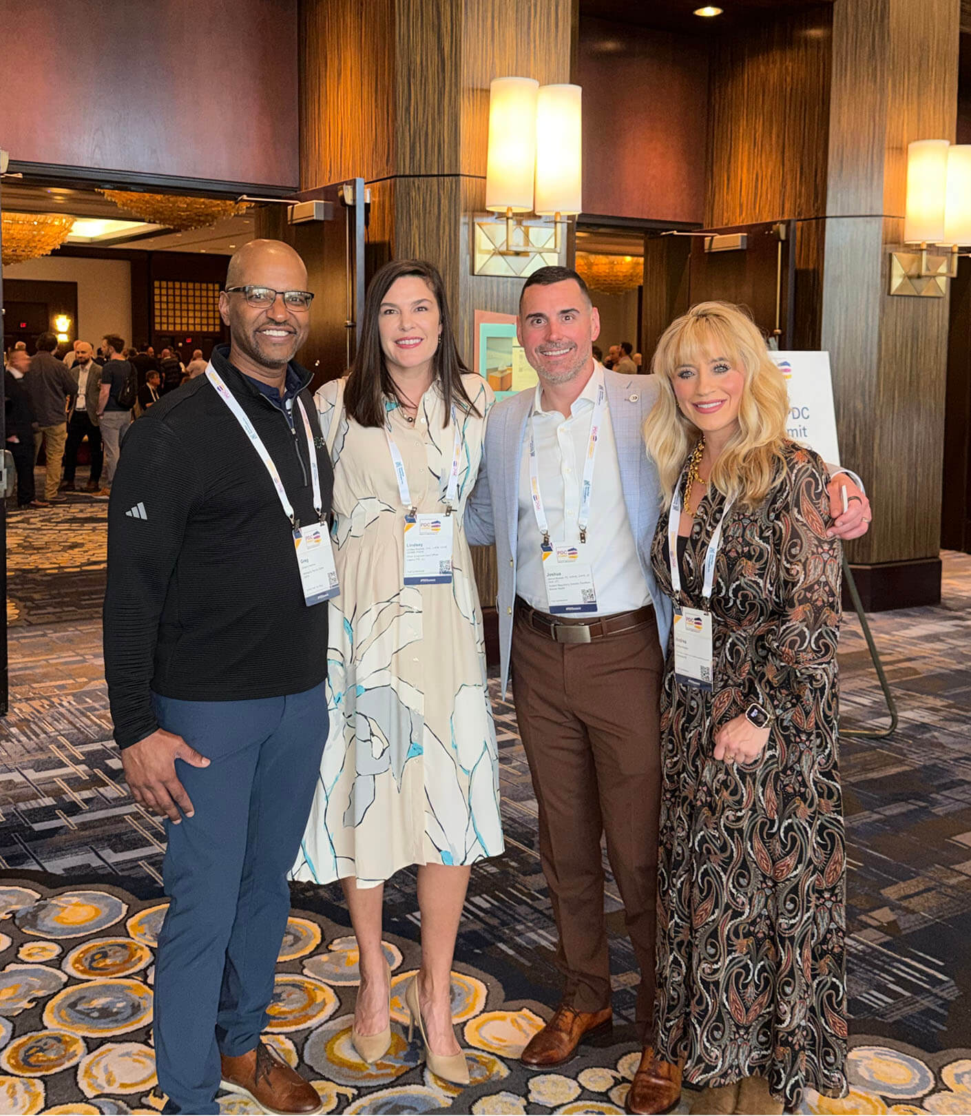 Andrea stands in a group of four with conference lanyards in a hotel lobby featuring wood paneling and decorative lighting.