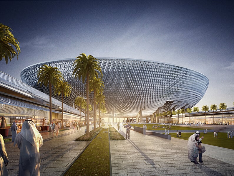 Large dome-shaped structure surrounded by palm trees, with people walking and sitting on a well-maintained pathway at sunset.