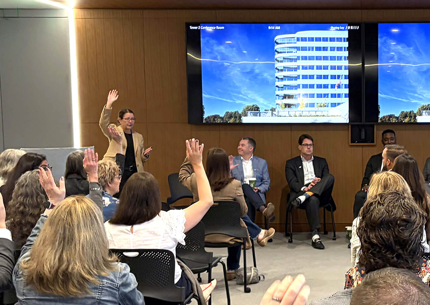 A speaker engaging an audience, with participants raising hands for questions. A large screen displays a building exterior.