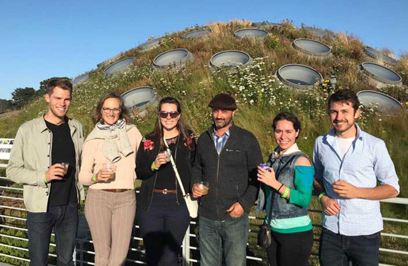 Group of six people in front of a green-roofed, domed structure with circular skylights, surrounded by wildflowers and grass.