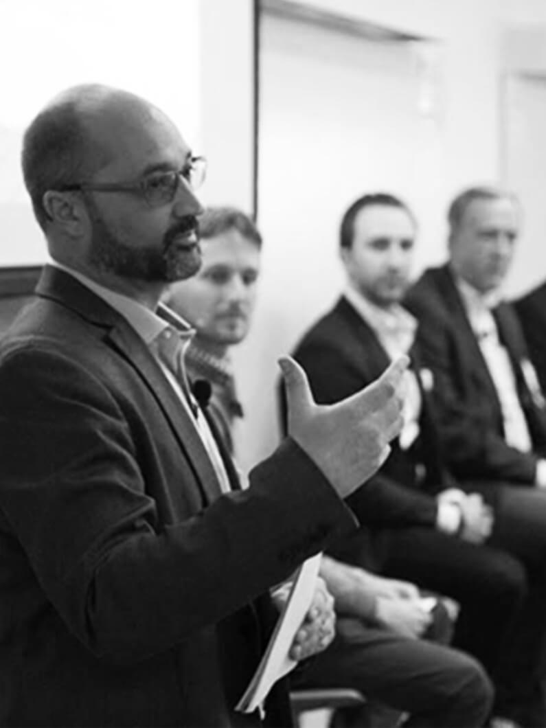 Speaker presenting in a formal setting, with a focus on architectural design concepts, while panelists listen intently in the background.