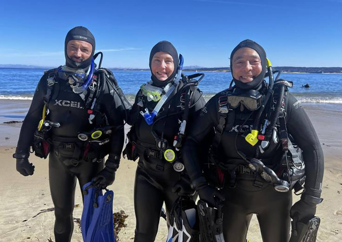 Three scuba divers in wetsuits stand on a sandy beach, ready to explore the ocean's depths, with calm water and distant hills in the background.