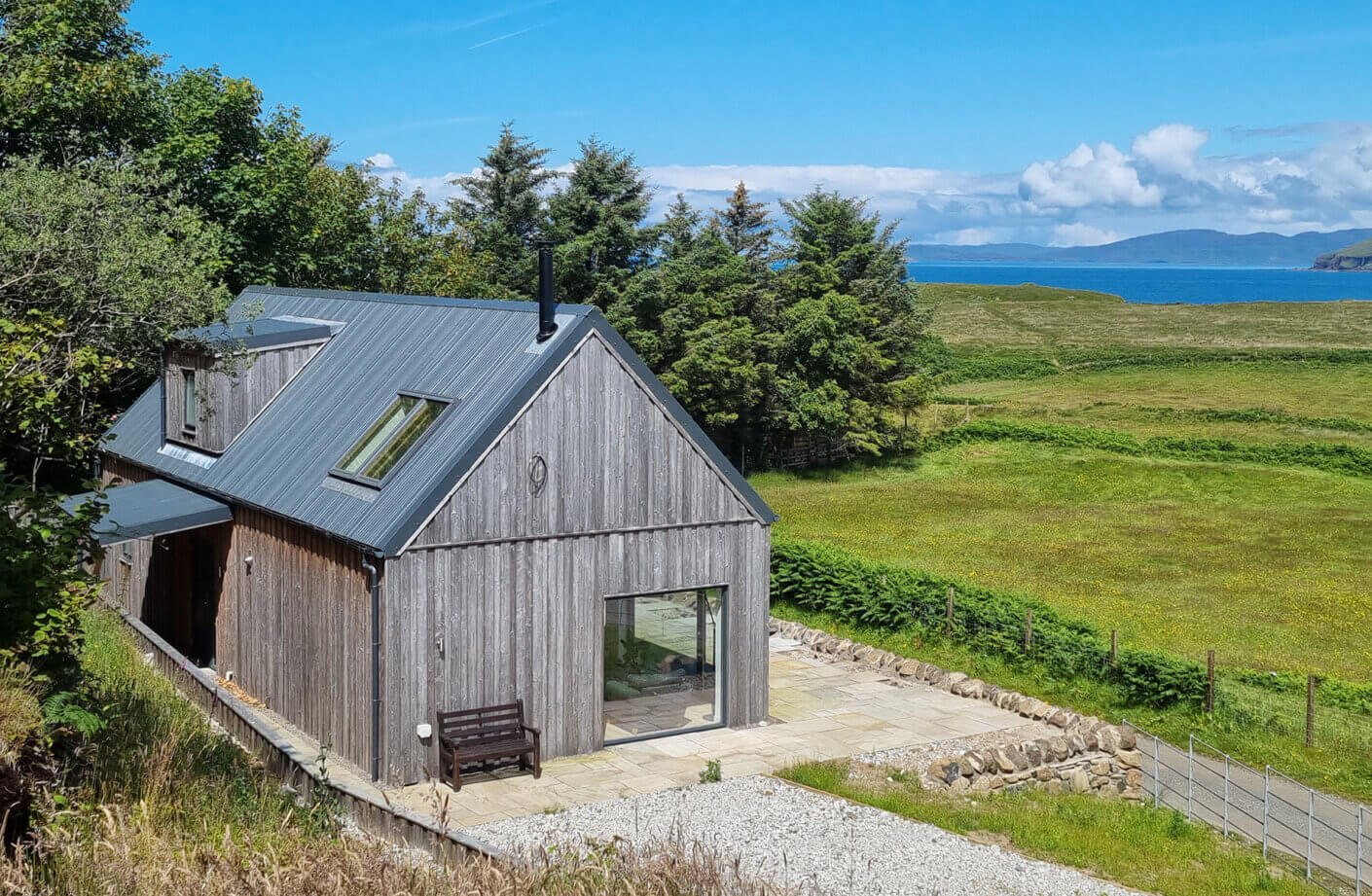 Wooden house featuring a metal roof, large windows, and a stone patio, set against a backdrop of trees and open landscape near the coast.