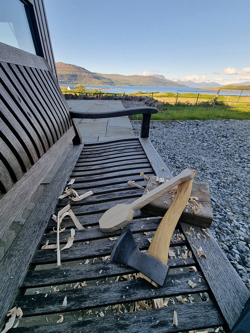 A wooden bench features a slatted design, complementing the rugged landscape. Nearby, carved utensils and tools.