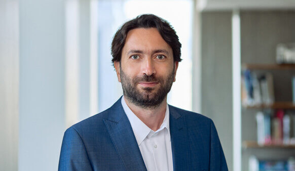 Professional man with dark hair and beard, wearing a suit, posed in a well-lit office setting with bookshelves in the background.