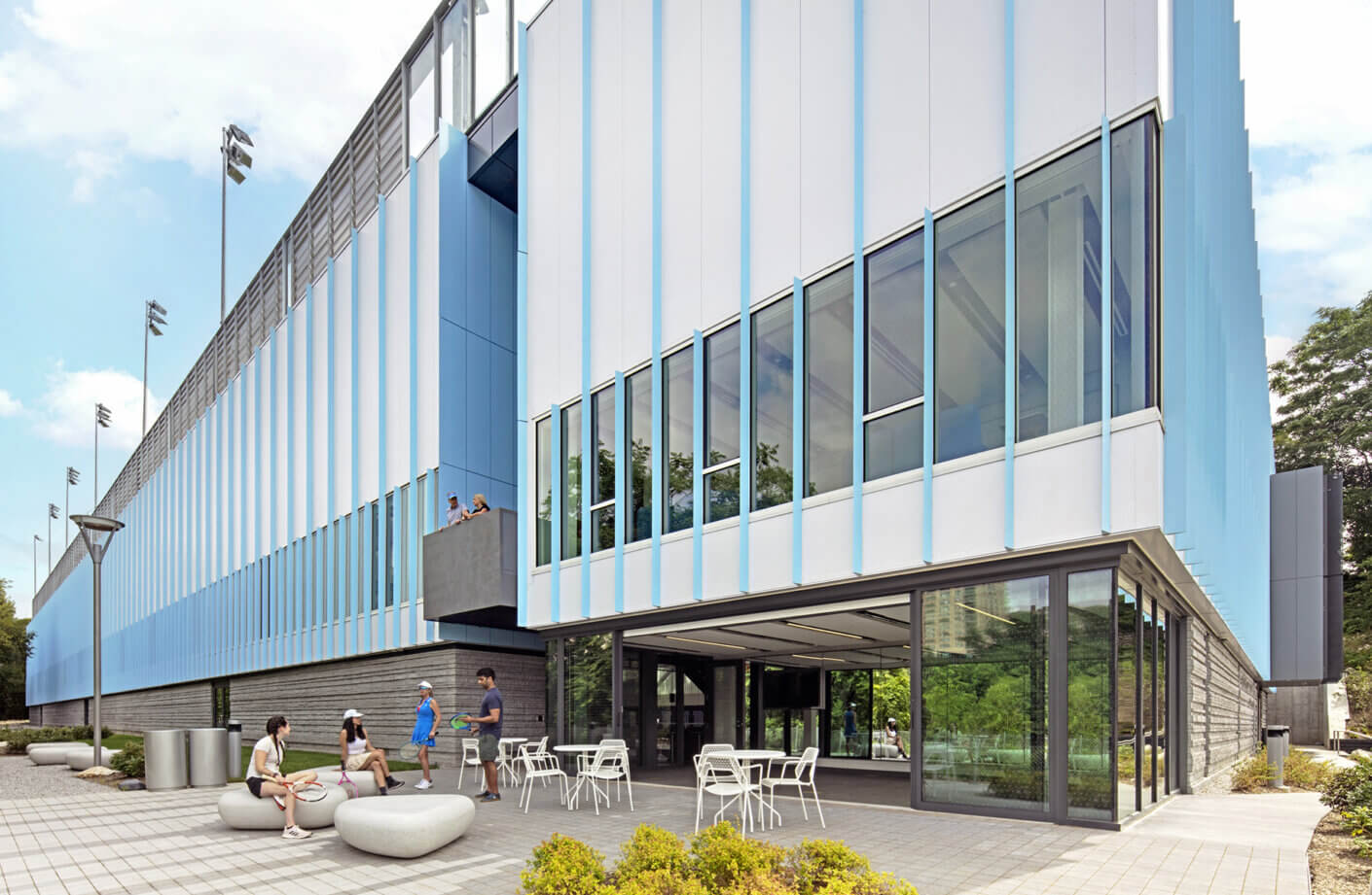 Image of a tennis facility with a sleek facade, featuring large windows, outdoor seating, and people engaging in activities outside.