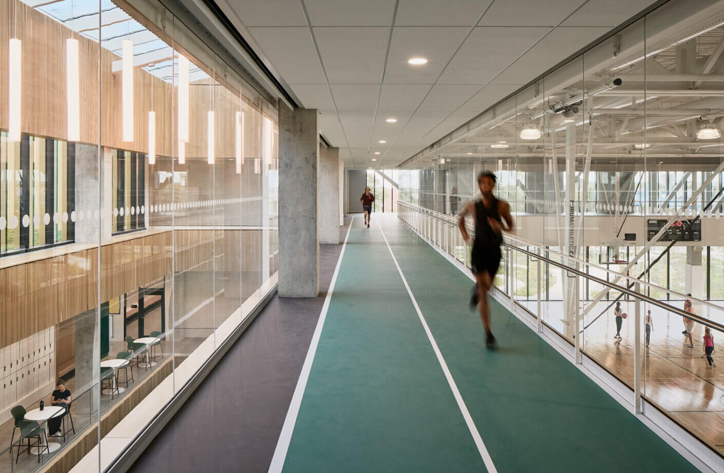 Two individuals running on an indoor track, with large windows and wood paneling visible. Tables and chairs are in the background.