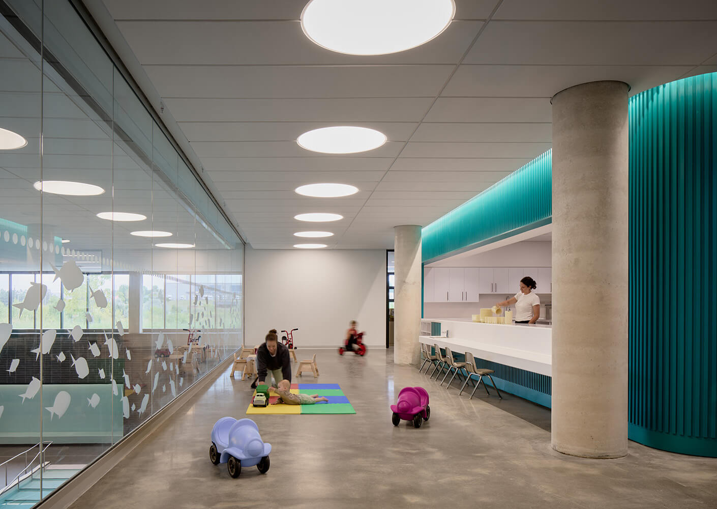 Interior view of a childcare space featuring play areas with toys, a seated adult at a counter, and large windows allowing natural light.