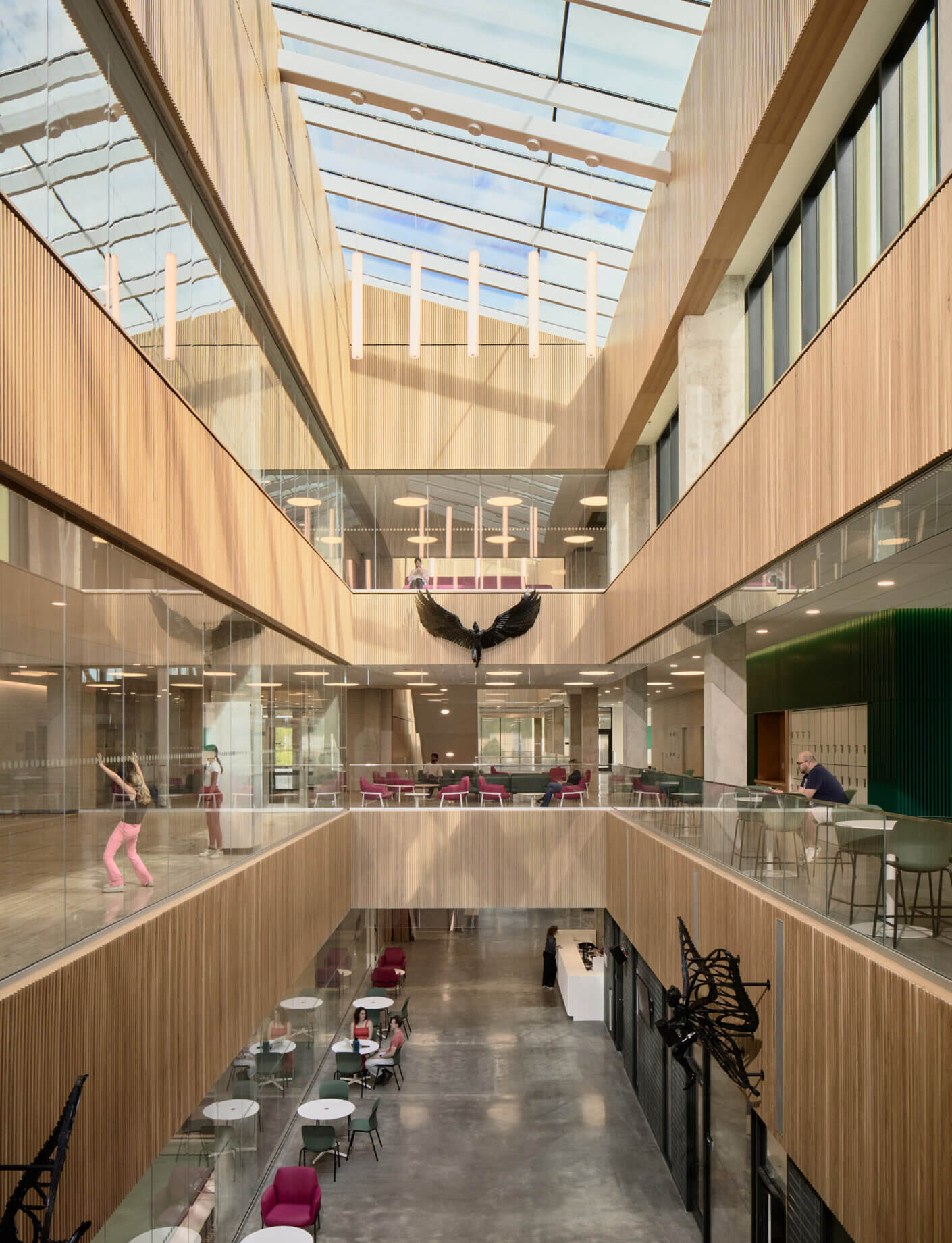 Interior view of an atrium with wooden paneling, glass railings, and multiple seating areas. People engage in activities, with art installations.