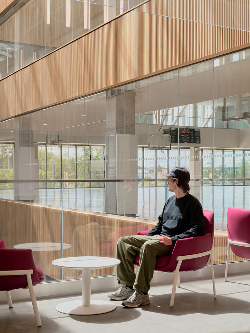 A person sits in a pink chair near a glass wall, overlooking a spacious interior with wood accents and natural light. A white table sits nearby.
