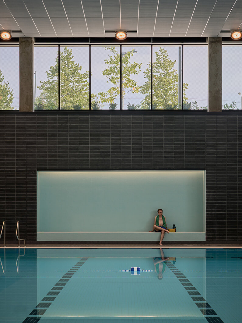 Indoor swimming pool in lower level with large windows letting in natural light, featuring a person sitting by the water's edge.