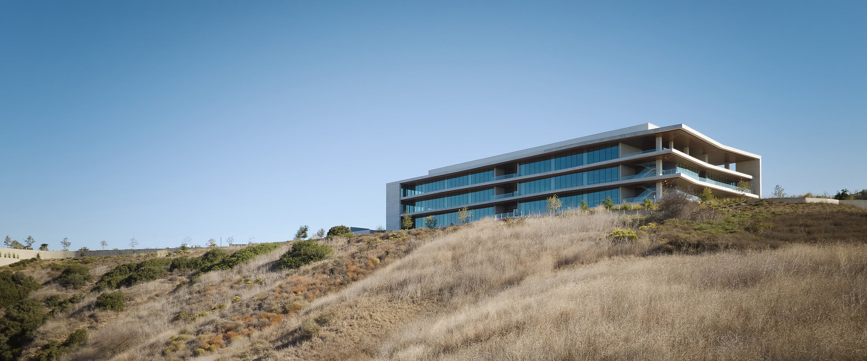 Contemporary building on a hillside, featuring large glass windows and multiple levels, surrounded by dry grass and sparse vegetation under a clear sky.