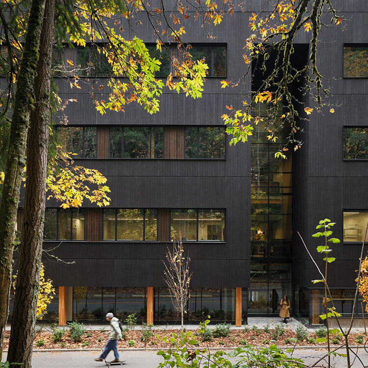 A pedestrian walks by a sleek, dark wooden building with large windows, set among autumn foliage and greenery. A second person is visible near the entrance.