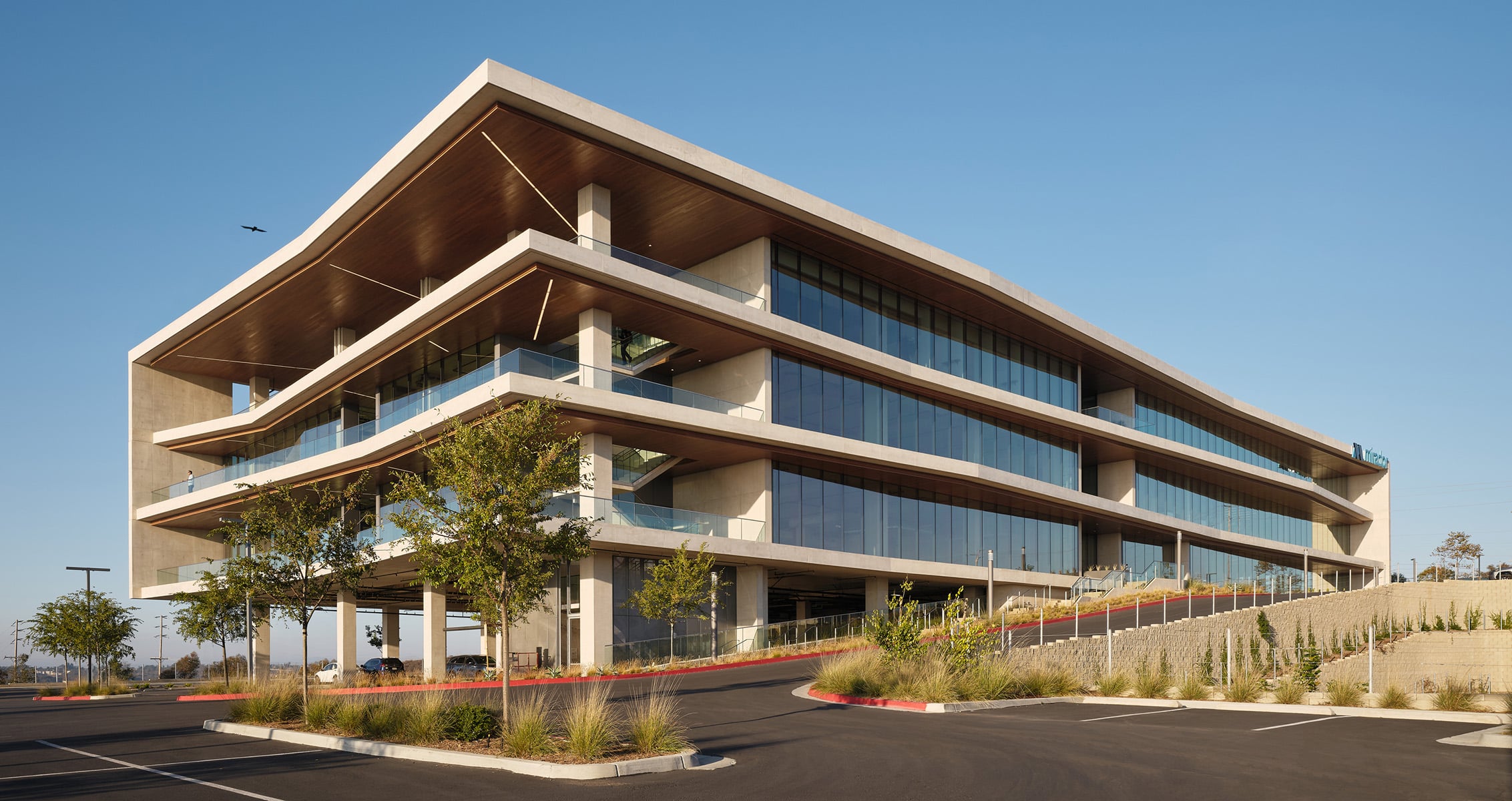 Contemporary office building with large glass windows, tiered balconies, and landscaped surroundings, set in a parking lot against a blue sky.