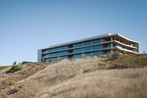 A hillside building with large glass windows, featuring multiple levels and a flat roof, surrounded by dry grass and sparse vegetation.