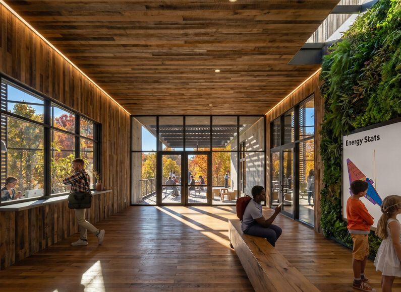 Inside view of a spacious building with large windows, wooden features, and a vertical garden, showing people engaged in various activities.