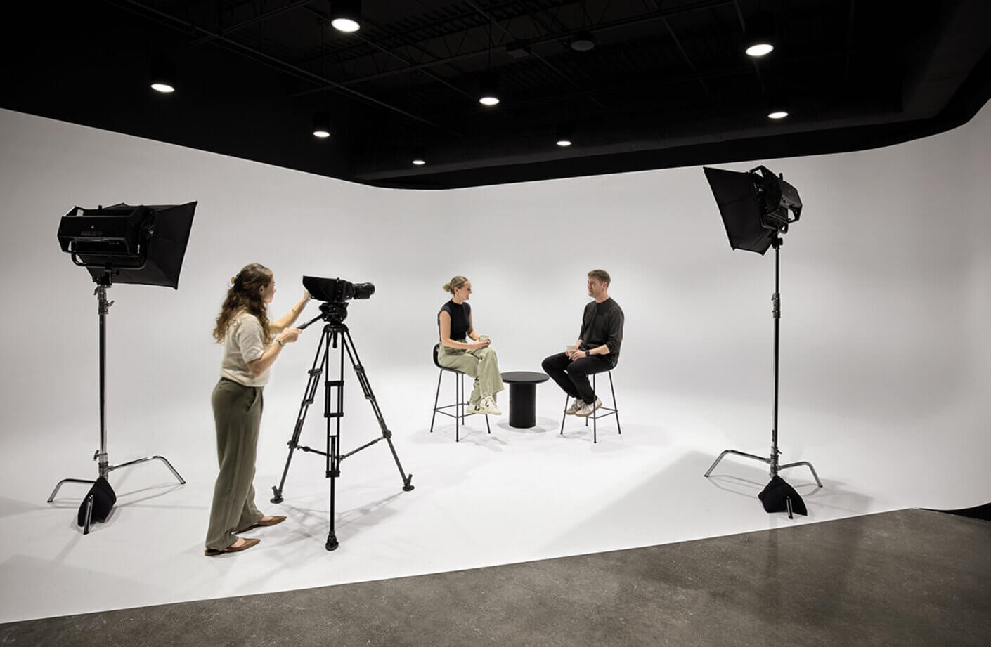 Two people seated for an interview on a minimalist set with a camera and lighting equipment in the background.