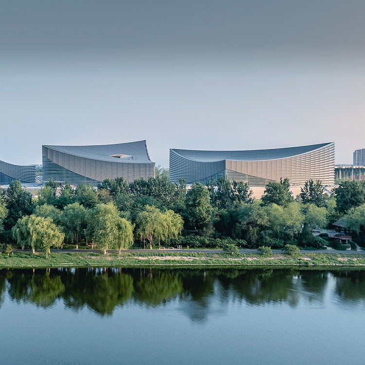Aerial view of three unique buildings next to a river, surrounded by trees reflecting in the water. Late afternoon light casts soft shadows.