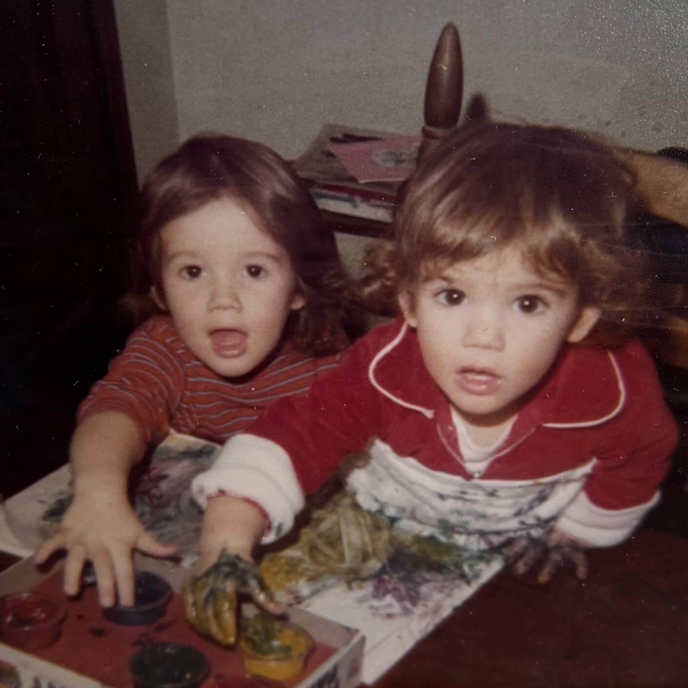 Two young children engaged in painting at a table, with colorful paint on their hands and the surface. One child is excited, while the other observes.