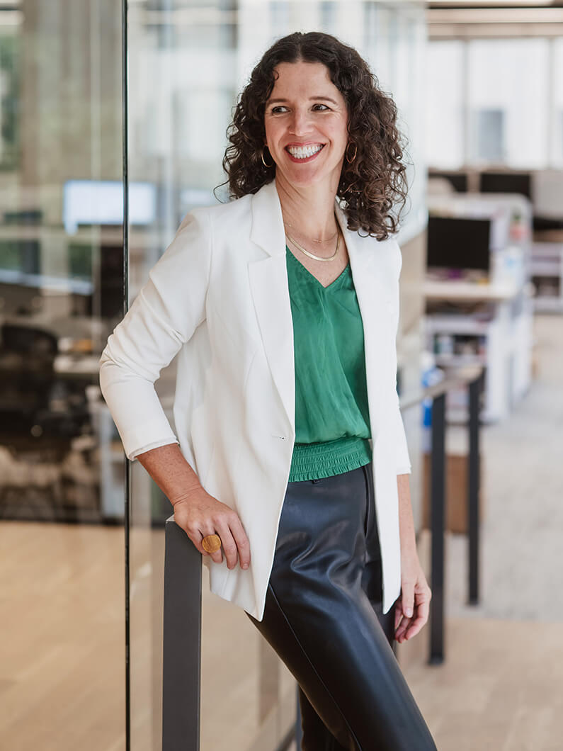 Smiling woman in a white blazer and green top leans against a glass partition in an office setting, showcasing a professional look.