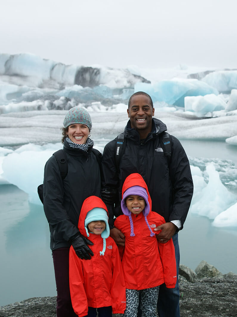 Family of four standing in front of a glacier, wearing colorful winter jackets. Icebergs visible in the background under a cloudy sky.