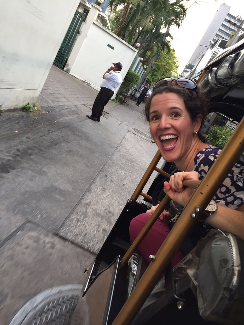 Tourist enjoying a ride in a tuk-tuk on a city street, with palm trees and buildings in the background. A man walks nearby.