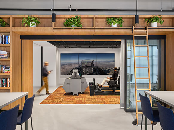 Interior office space featuring seating areas, large screens, and greenery. A person walks by while another is seated at a control station.