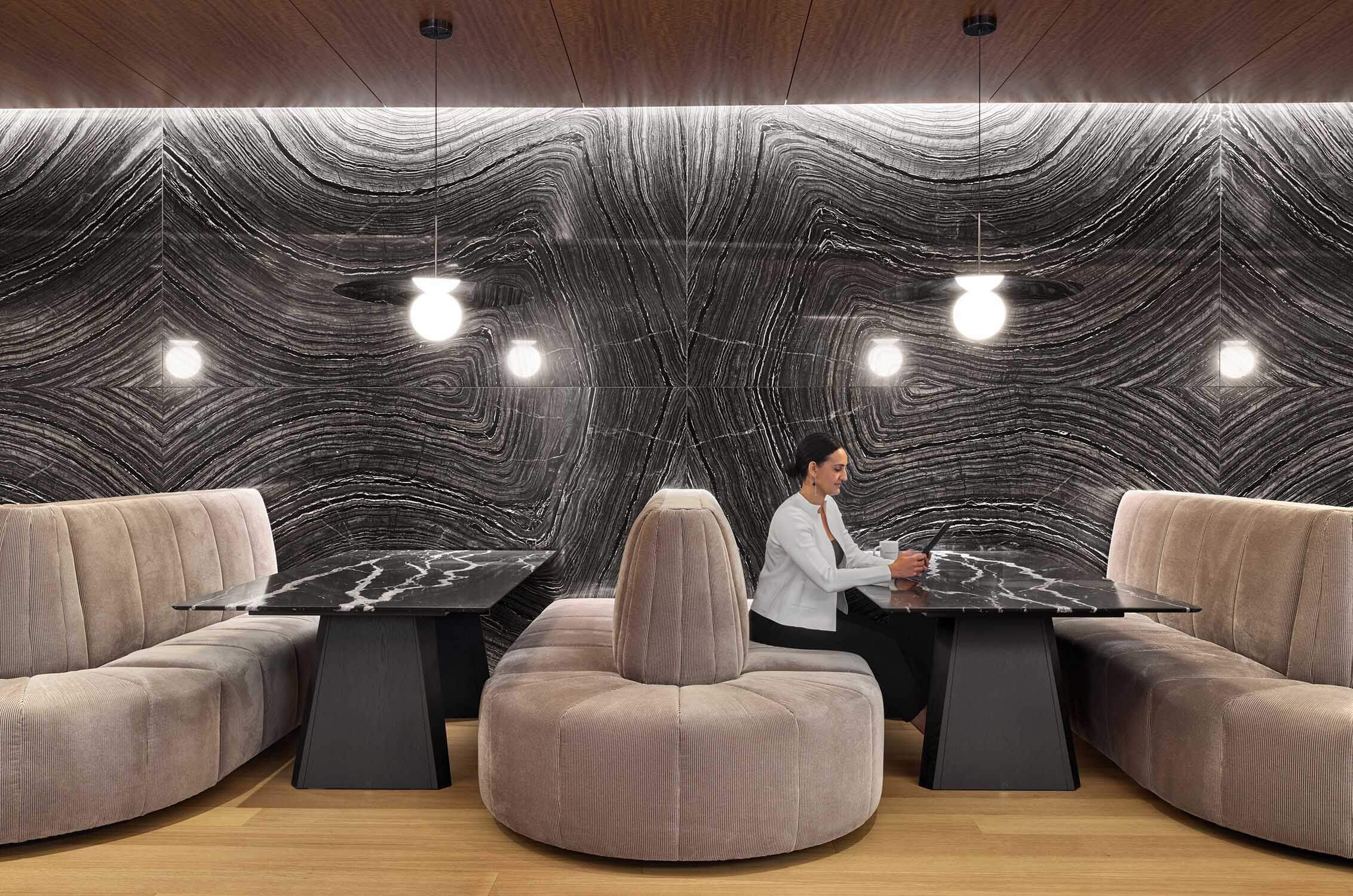 Woman sitting at a sleek table in a stylish seating area, with a textured black wall and hanging lights in the background.