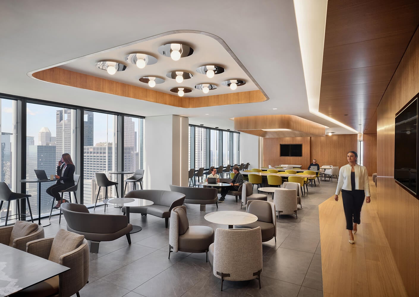 Office dining lounge with large windows, contemporary seating, and a mix of workspaces. People engaged in conversation and using laptops.