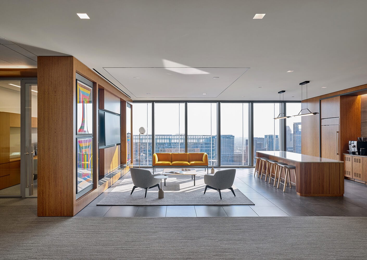 Bright office interior featuring a yellow sofa, gray armchairs, a wooden bar counter, and large windows overlooking the city skyline.
