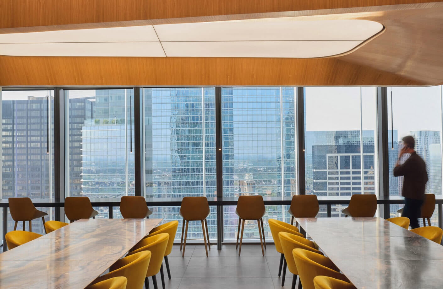 Office dining lounge with long tables and yellow chairs, featuring floor-to-ceiling windows showing a city skyline and a person walking by.