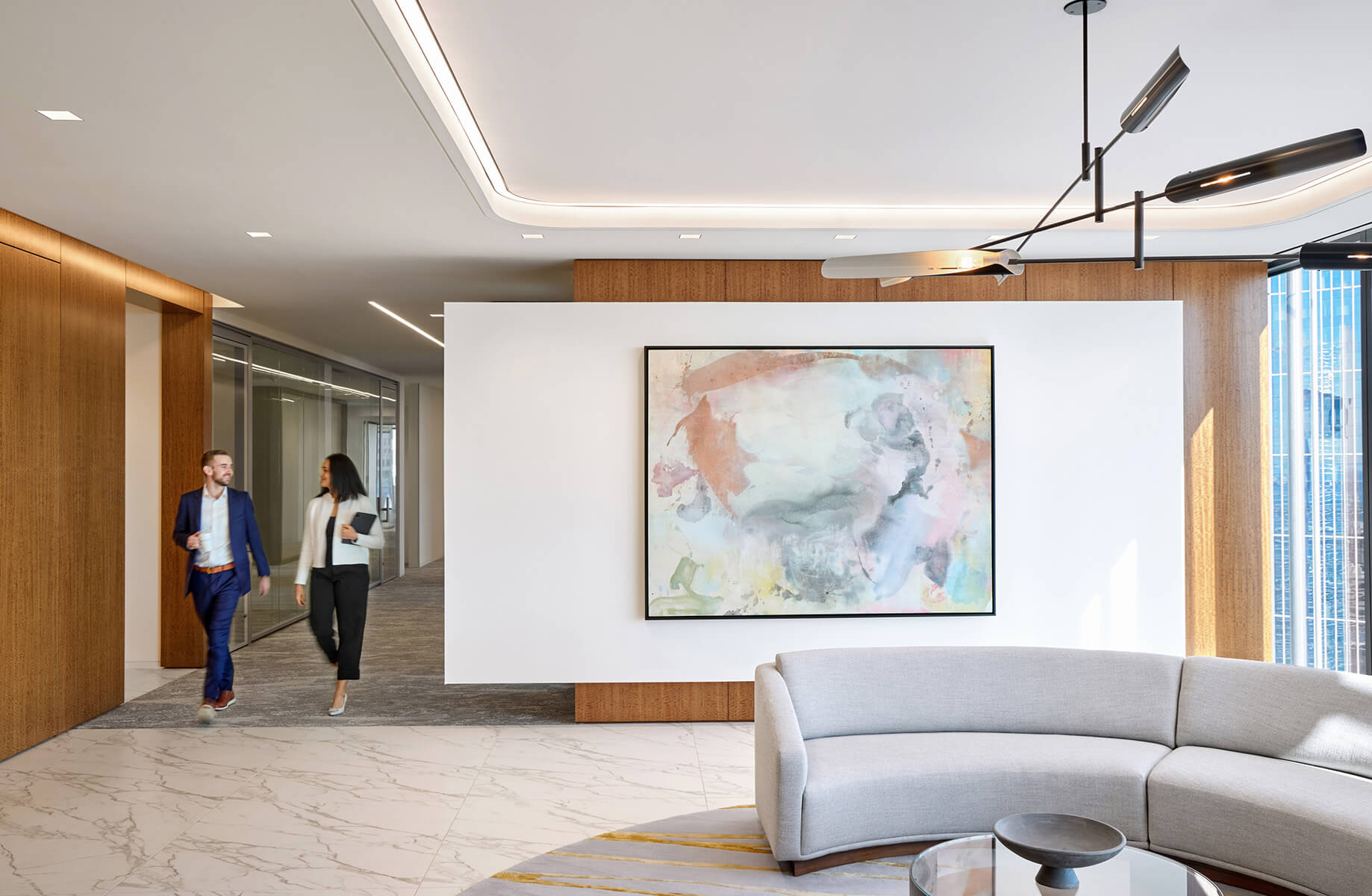 Two people walk in a bright office corridor, featuring artwork on the wall and a curved sofa in the foreground. Natural light streams through windows.