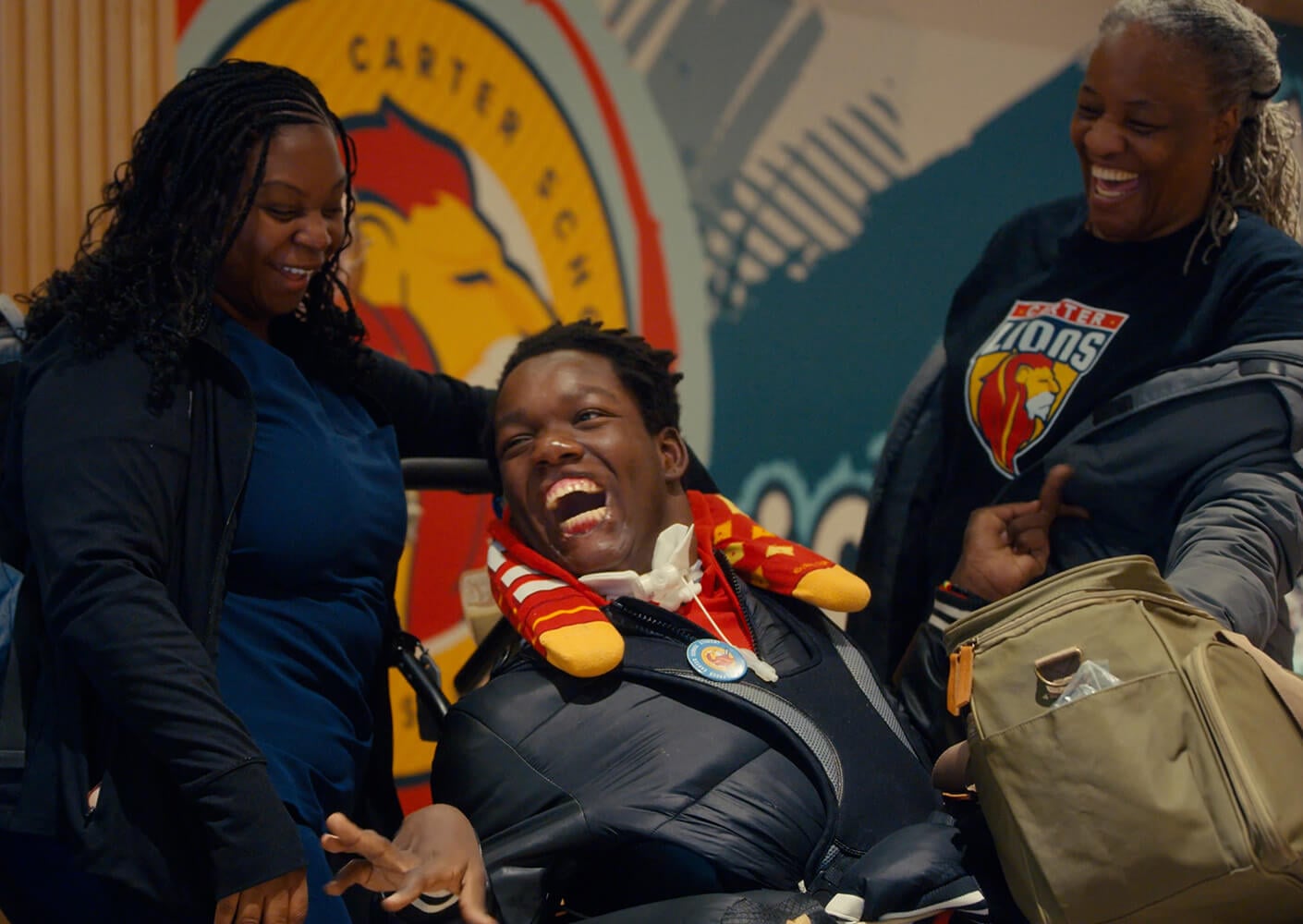 A joyful moment featuring two women and a young man in a wheelchair, with a school logo in the background and laughter shared among them.