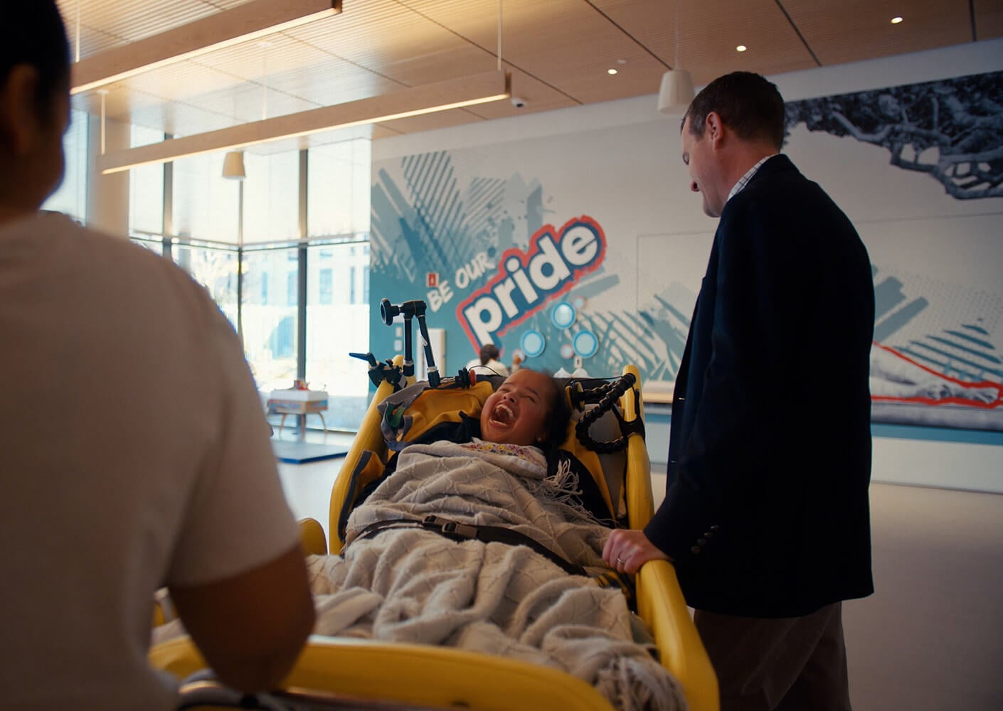 Child in a stretcher smiles at an adult, with a colorful wall mural in the background that reads Be Our Pride.