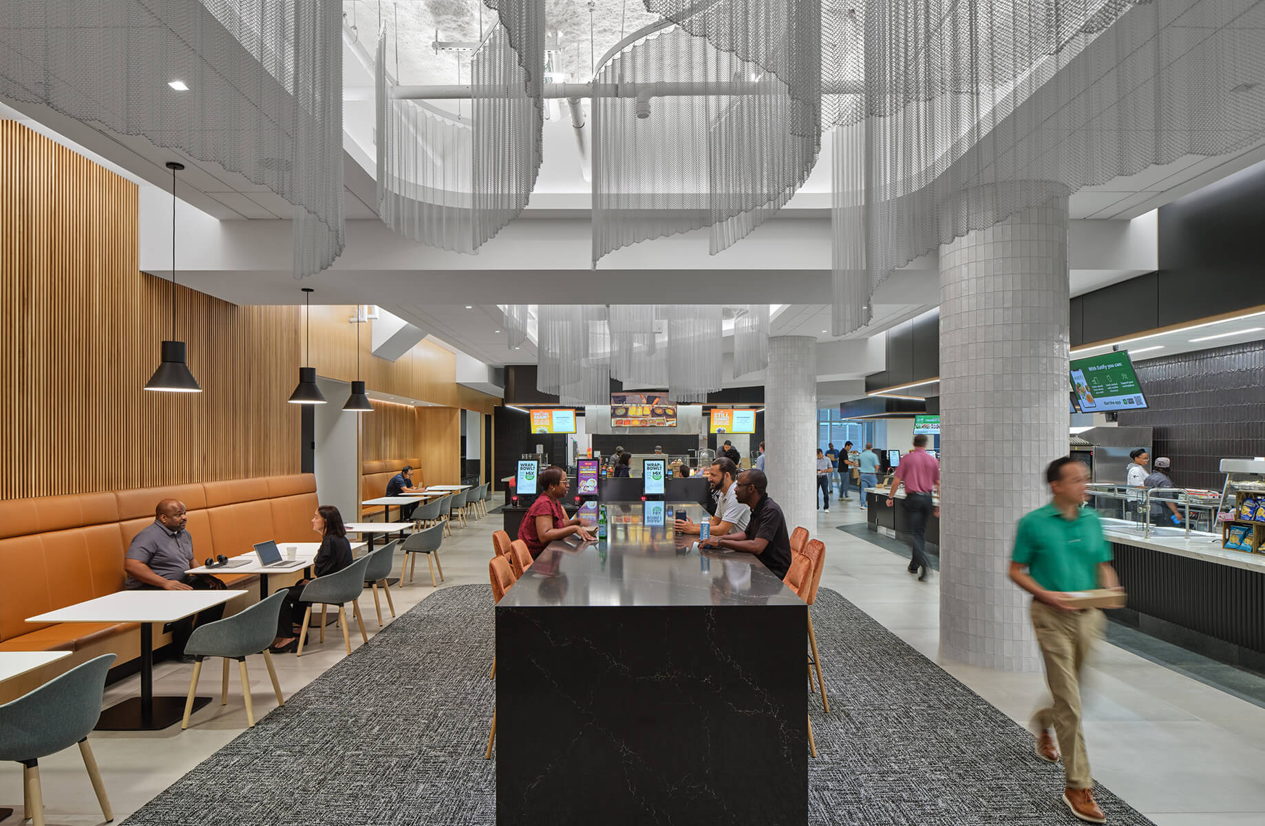 Casual dining space with varied seating, hanging fixtures, and a service area. People engage at tables and a buffet. Natural light from windows.
